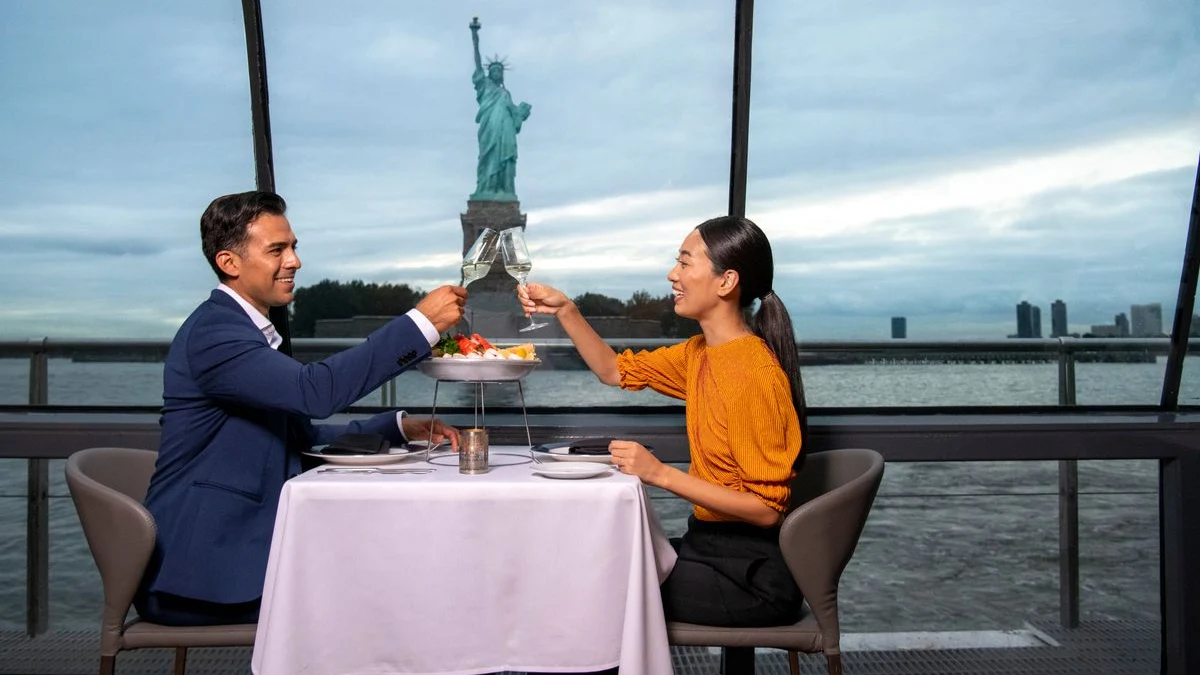 A couple toasting champagne in the Brunch Cruise with the Liberty statue in the backdrop